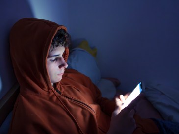 Sad and Tired Teenager Lying in Bed at Night Using Smartphone.  Teenager using a smartphone lying in bed late at night, playing games, watching videos online, and scrolling the screen. Children's screen addiction. Screen Addiction in Youth